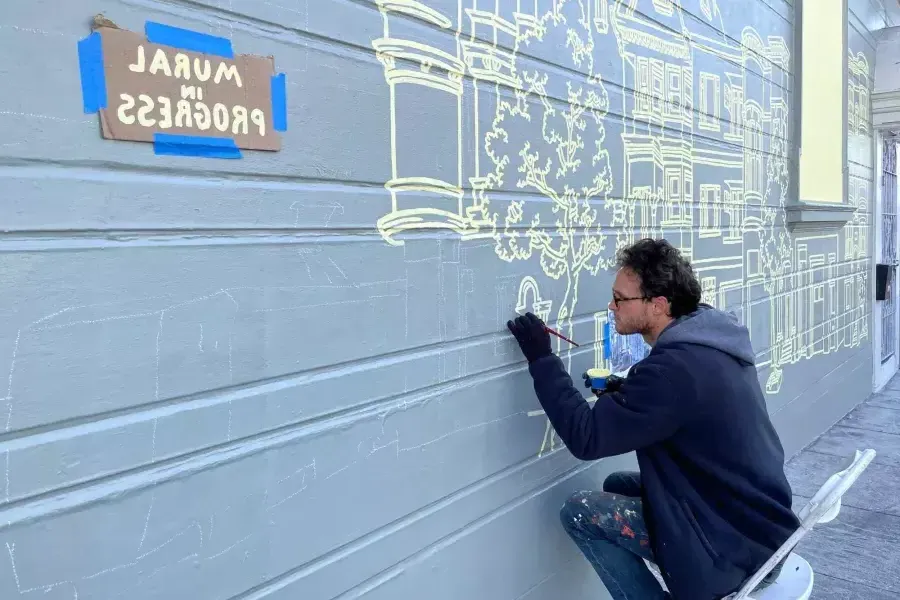 An artist paints a mural on the side of a building in the Mission District, with a sign taped onto the building that reads "Mural in Progress.“是贝博体彩app,ca.